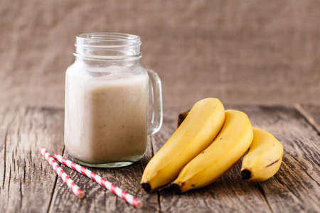 Delicious banana smoothie in glass jar with drinking straw and bananas on table.の写真素材