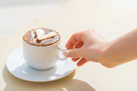 Girl take a white cup of coffee with milk and decorative heart on a deck chair near the pool....
