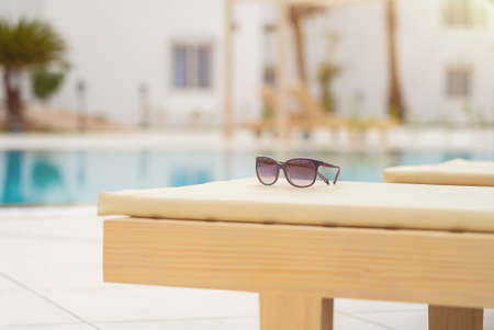 Close-up of woman sunglasses on deck chair near the pool on a bright sunny day. Resort vacation...