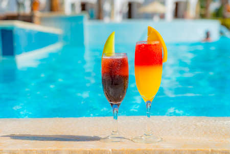 Two cocktails with natural juice and ice at the edge of the swimming pool. Orange cocktail with an...
