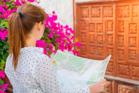 Girl holds a map in her hands, under the bush with flowers and in front of wooden door. Street of...