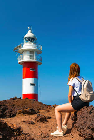 Young woman with backpack sit in front of a lighthouse on a rocky shore. Girl near a phare on a voulcanic rocks, sunny day with bright blue sky. Beacon on a oceanic shoreの写真素材