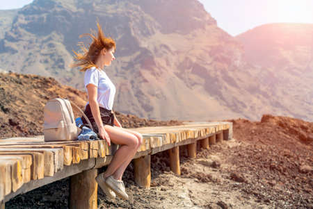 Young woman with backpack sits on wooden platform on a rocky ocean shore. Happy girl on a sunny day with bright blue sky. Voulcanic mountains of Tenerife on backgroundの写真素材