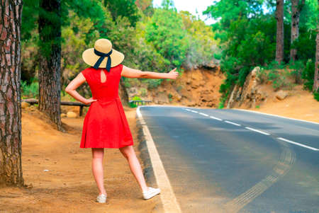 Woman in red dress by the road in forest trying to hitchhike a rideの写真素材