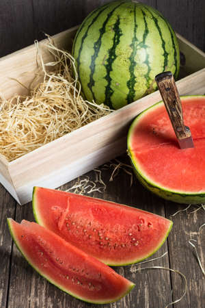 A whole watermelon in a wooden box and slices of ripe watermelon, a knife with a wooden handle on a wooden background. View from above.の写真素材