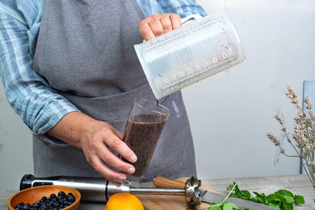 woman pours prepared healthy detox drink in a blender into a glass - a green smoothie with fresh fruits, blueberries, green spinach and kiwi.の写真素材