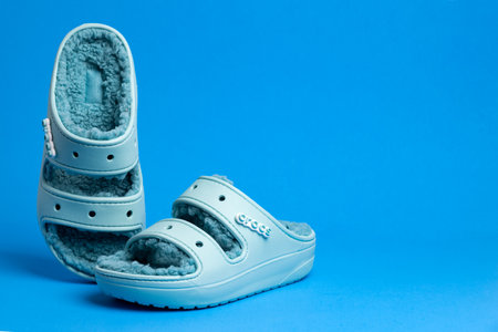 Turquoise Crocs rubber slippers trimmed with faux fur. Comfortable, light shoes on a blue background.のeditorial素材