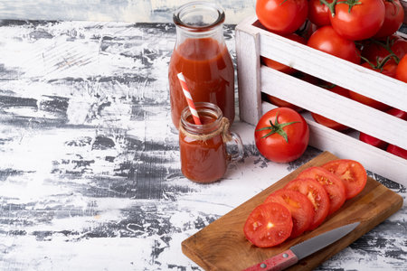Ripe fresh tomatoes in a wooden box, tomato slices on a cutting board and a bottle of tomato juice on a white table.の写真素材