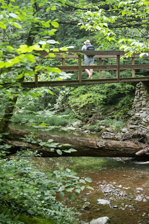 woman tourist in a hat with a backpack stands on a wooden bridge across a mountain river in the...