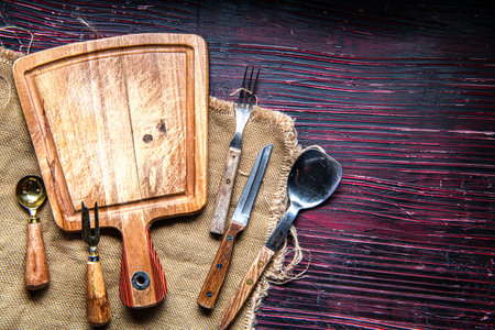Cutting board, forks, spoons and knives on a dark red wooden table. Space for text.の写真素材