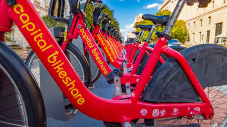 Row of red public bikeshare bicycles parked at a docking station on a sunny spring day near a city park and pedestrian crossing.のeditorial素材