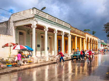 A busy street scene in Cuba after a tropical downpour showing people cycling and shopping.