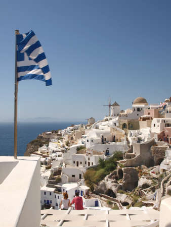 A couple looking at a view of a collection of holiday accommodations and windmills in Santorini, showing a Greek flag blowing in the breezeの写真素材