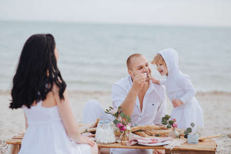 Daddy feeds the child at a table near the sea, mom sits back. Family sea tourの写真素材