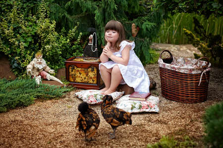 Little girl in white dress sitting in the garden and watching the chickensの写真素材