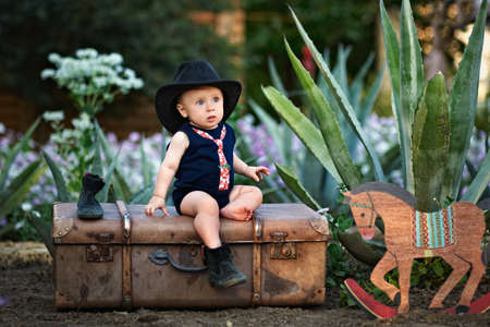 Little boy in a black hat sitting on a vintage suitcase in the gardenの写真素材