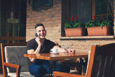 Guy sitting at a table in a cafe in the street, talking on the phone and smiling. On the table is a cup of coffee and a glass of waterの写真素材