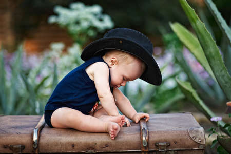 Little boy in a black hat sitting on a vintage suitcase in the gardenの写真素材