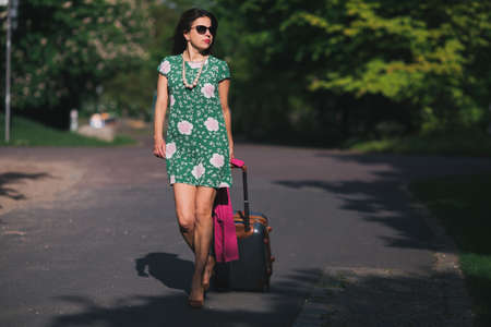 Girl in a dress and wearing spectacles goes along the road with a suitcase among green trees. Concept of a travelの写真素材