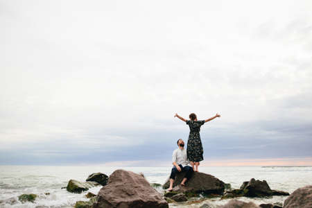 happy couple in love on the beach. man sitting on the rocks and woman standing with open handsの写真素材