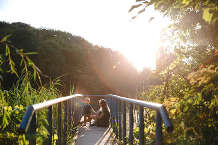 Front view mother and baby son sitting on the bridge on lake in summer evening timeの写真素材