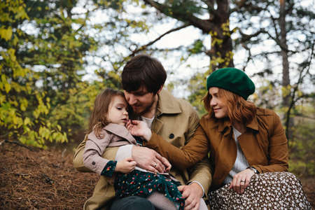 Young father, mother and little toddler daughter girl in a beret and a coat sitting on the ground in...