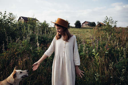 Beautiful young woman relaxed and carefree enjoying a summer sunset with her lovely dog on the straw and high flowers field backgroundの写真素材