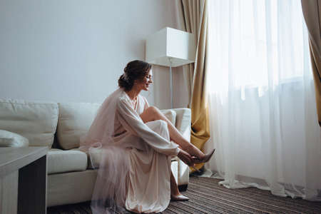 beautiful bride is preparing for a meeting with her fiance in the white room. Girl in wedding dress in front of the window putting on high-heeled shoesの写真素材