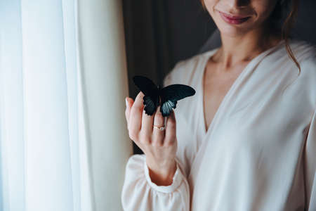 A beautiful gentle bride in a beige peignoir with a butterfly with black and blue wings sits on the hand of the bride, she smilesの写真素材