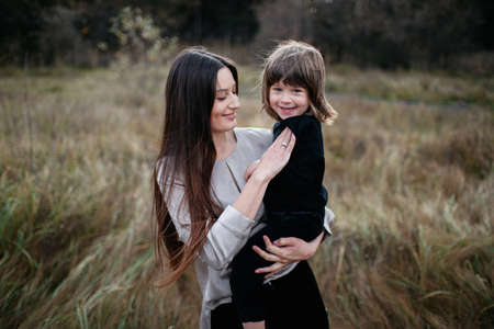 woman with son in her arms on dry grass field and forest background. Happy mom with long dark hair and her sonの写真素材