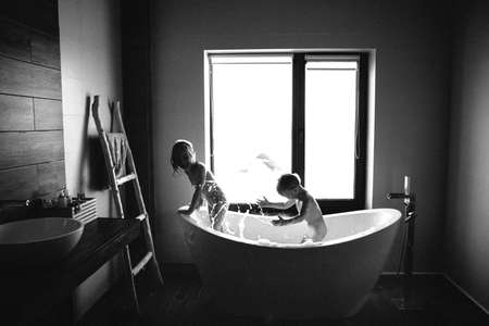 Black and white photo of brother and sister splashing in the bathroom. Side view. Oval bath indoor. Happy and healthy childhood conceptの写真素材