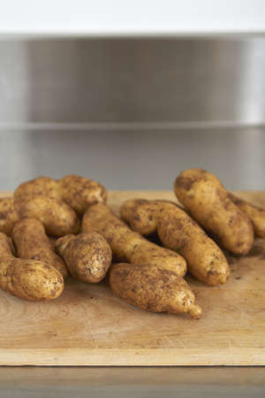Wooden chopping board with fresh, dirty potatoes. Stainless steel bench in background. Shallow focusの写真素材
