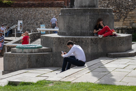 09/02/2019 Winchester, Hampshire, UK A man in business clothing sitting on the steps of a war memorial using his mobile phone all cell phone surrounded by people relaxing in the parkのeditorial素材
