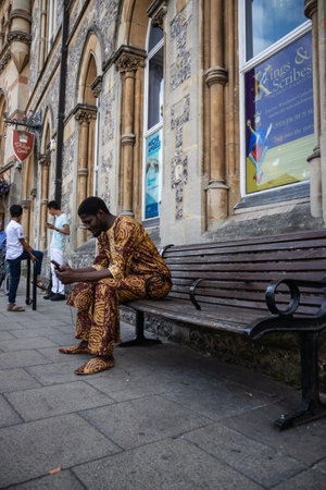 09/02/2019 Winchester, Hampshire, UK a black man wearing traditional african dress sitting on a bench looking at his mobile phone or cell phoneのeditorial素材