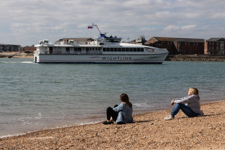09/14/2019 Portsmouth, Hampshire, UK two women sitting together on a pebble beach in the summer with a ferry passingのeditorial素材