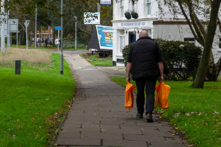 11/05/2019 Portsmouth, Hampshire, UK a man walking alone carrying two orange shopping bags homeのeditorial素材