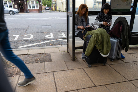 11/06/2019 Winchester, Hampshire, UK two asian women sitting at a bus stop with suitcases while people walk byのeditorial素材