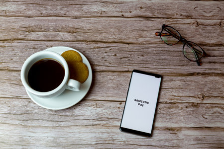 01-30-2021 Portsmouth, Hampshire, UK A Mobile phone or cell phone laid on a wooden table with the samsung pay app opening also a coffee and glassesのeditorial素材