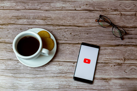 01-30-2021 Portsmouth, Hampshire, UK A Mobile phone or cell phone laid on a wooden table with the Youtube app opening also a coffee and glassesのeditorial素材