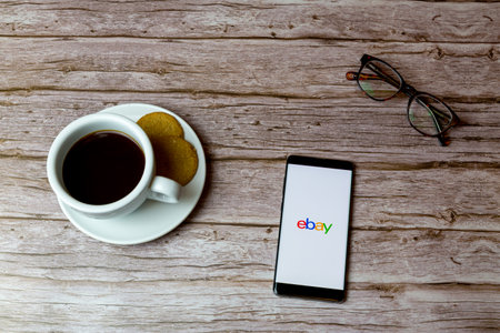 01-30-2021 Portsmouth, Hampshire, UK A Mobile phone or cell phone laid on a wooden table with an Ebay app opening also a coffee and glassesのeditorial素材