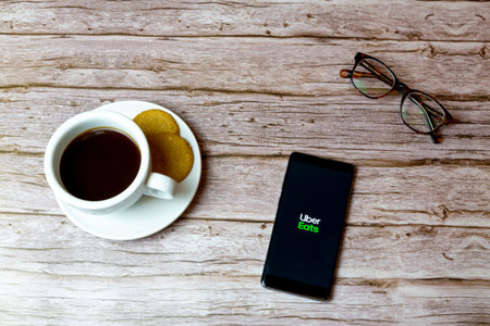 01-30-2021 Portsmouth, Hampshire, UK A Mobile phone or cell phone laid on a wooden table with Uber eats app opening also a coffee and glassesのeditorial素材