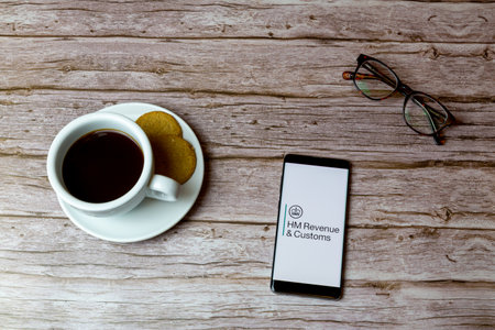 01-30-2021 Portsmouth, Hampshire, UK A Mobile phone or cell phone laid on a wooden table with an HMRC app opening also a coffee and glassesのeditorial素材