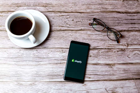 02-08-2021 Portsmouth, Hampshire, UK A mobile phone or cell phone laid on a wooden table with the shopify app open next to a coffee and glassesのeditorial素材