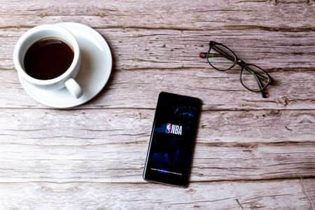 02-08-2021 Portsmouth, Hampshire, UK A mobile phone or cell phone laid on a wooden table with the NBA app open next to a coffee and glassesのeditorial素材