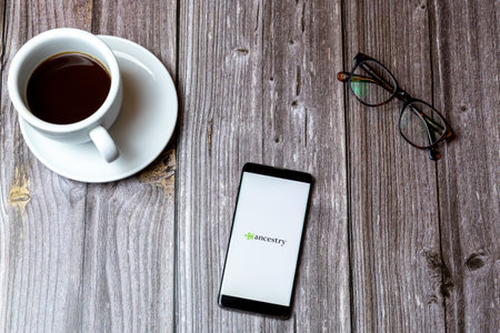 02/10/2021 Portsmouth, Hampshire, UK A mobile phone or cell phone laid on a wooden table with the Ancestry app open on screen next to a coffeeのeditorial素材