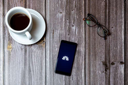 02/10/2021 Portsmouth, Hampshire, UK A mobile phone or cell phone laid on a wooden table with the NBC News app open on screen next to a coffeeのeditorial素材
