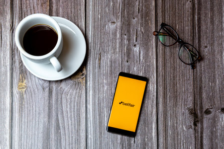 02/10/2021 Portsmouth, Hampshire, UK A mobile phone or cell phone laid on a wooden table with the Betfair betting app open on screen next to a coffeeのeditorial素材
