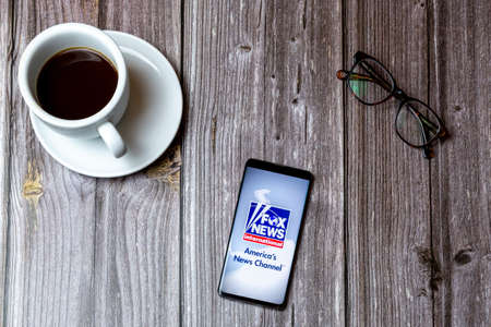 02/10/2021 Portsmouth, Hampshire, UK A mobile phone or cell phone laid on a wooden table with the Fox News app open on screen next to a coffeeのeditorial素材