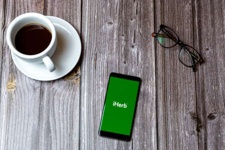 02/10/2021 Portsmouth, Hampshire, UK A mobile phone or cell phone laid on a wooden table with the iHerb app open on screen next to a coffeeのeditorial素材