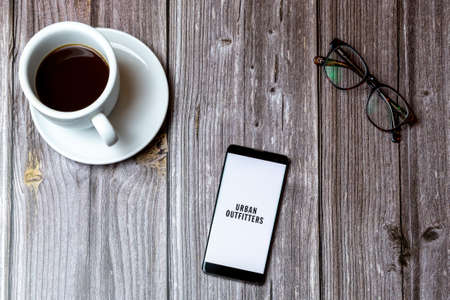 02/10/2021 Portsmouth, Hampshire, UK A mobile phone or cell phone laid on a wooden table with the Urban Outfitters app open on screen next to a coffeeのeditorial素材